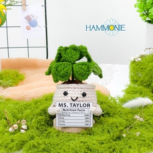 May include: A crocheted green four-leaf clover plant in a white pot with a smiling face. The pot has a white label that reads "MS. TAYLOR Nutrition Facts" with a list of qualities and percentages. The plant is sitting on a bed of green moss.
