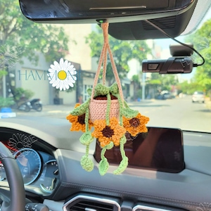 May include: A crocheted sunflower hanging from the rearview mirror of a car. The sunflower is in a small pot and has green leaves. The sunflower is yellow and brown.