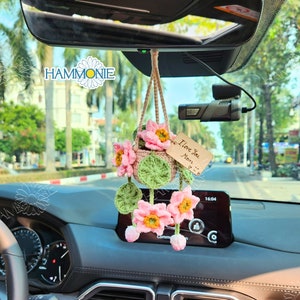 May include: A crocheted flower arrangement hanging from the rearview mirror of a car. The flowers are pink and white, and the arrangement includes a small green basket. A wooden tag with the text "I love you Mom" is attached to the arrangement.
