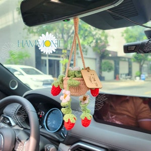 May include: A crocheted hanging planter with strawberries and white flowers. The planter is hanging from the rearview mirror of a car. A wooden tag with the text "I love you Mom!" is attached to the planter.