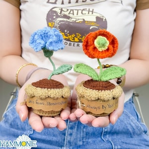 May include: Two crocheted potted plants, one with a blue flower and the other with an orange flower. The pots have brown bases and beige crocheted edges. The pots have labels that read "Love is Sown, Memories Bloom" and "Always By Your Side".