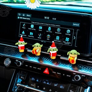 May include: Four green and brown crocheted Yoda ornaments wearing Santa hats and holding gifts. The ornaments are hanging from the dashboard of a car.