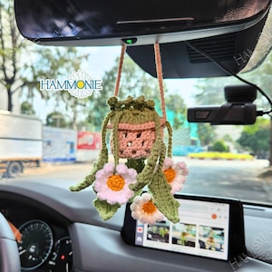 May include: A crocheted hanging decoration featuring a small pink pot with green leaves and white flowers. The decoration is hanging from the rearview mirror of a car. The text "HAMMONIE" is visible in the background.