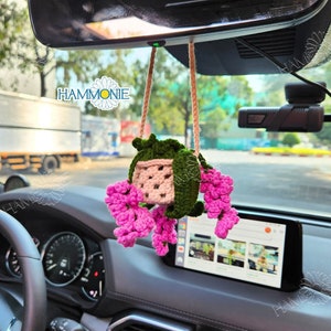May include: A pink and green crocheted hanging flower basket with a small white pot inside. The basket is hanging from the rear-view mirror of a car.