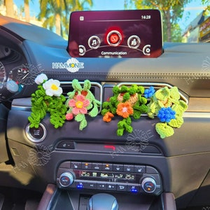 May include: Four crocheted flower arrangements in different colours, including pink, orange, blue, and white, are displayed in the air vents of a car's dashboard. The flowers are made with yarn and have a realistic appearance.