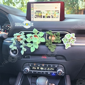 May include: Four crocheted green plant air fresheners hanging from the air vents in a car. The plants are in small brown pots and have green leaves. The car's dashboard is visible in the background.