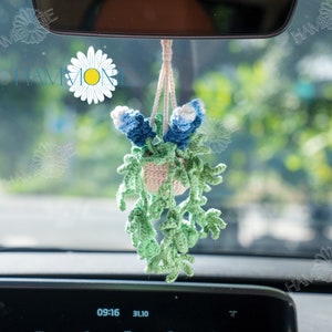May include: A crocheted hanging plant with blue flowers and green leaves. The plant is hanging from the rearview mirror of a car.