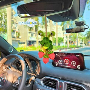 May include: A green and red crocheted strawberry and snail car charm hanging from the rearview mirror of a car. The charm is handmade and features a cute snail with googly eyes and a bunch of strawberries.