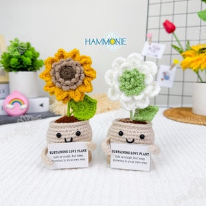 May include: Two crocheted potted plants with happy faces. The sunflower plant has a yellow and brown flower and green leaves. The other plant has a white and green flower and green leaves. Both plants have a sign that reads "Sustaining Love Plant. Life is tough, but keep growing in your own way."