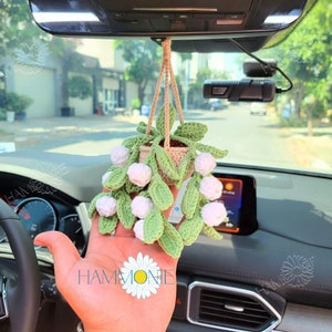 May include: A crocheted hanging plant with white flowers and green leaves. The plant is hanging from the rearview mirror of a car.