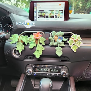 May include: Crocheted car vent decorations shaped like flowers and leaves. The flowers are in shades of orange, blue, and white, with green leaves. The decorations are displayed on a car dashboard.