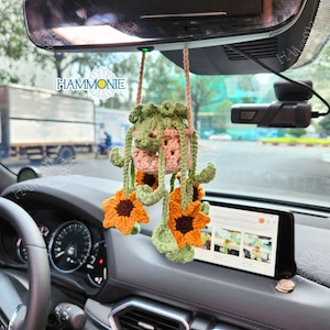 May include: A crocheted car air freshener with a pink and green hanging planter and four yellow sunflowers. The air freshener is hanging from the rear view mirror of a car.