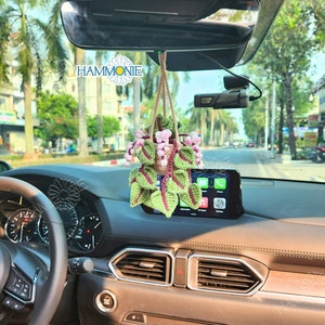 May include: A crocheted hanging plant with green leaves and pink flowers in a brown pot. The plant is hanging from the rearview mirror of a car. The text "HAMMONIE" is visible in the upper left corner of the image.
