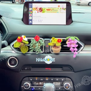May include: Four crocheted flower pot air fresheners hanging from the air vents of a car. The air fresheners are in different colours and feature different types of flowers. The car's dashboard is visible in the background.