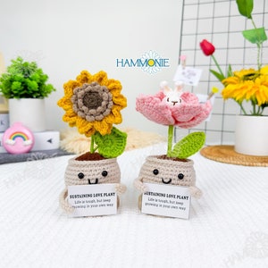 May include: Two crocheted potted plants with happy faces. The first plant has a yellow sunflower and green leaves. The second plant has a pink flower with a white bunny in the center. Both plants have a sign that reads "Sustaining Love Plant. Life is tough, but keep growing in your own way."
