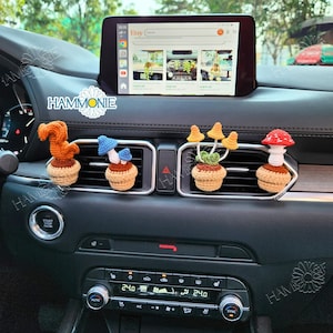May include: Four crocheted car air freshener ornaments in a car. The ornaments are a squirrel, a mushroom, a mushroom cluster, and a mushroom with a red cap. The ornaments are hanging from the air vents in the car.