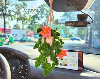 May include: A crocheted hanging plant with pink flowers in a white pot, hanging from the rearview mirror of a car. The plant is green and has a string of leaves hanging down. The car is black and has a dashboard and steering wheel visible.