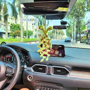 May include: A green crocheted plant-like figure with two eyes hangs from the rearview mirror of a car. The figure is a car charm.