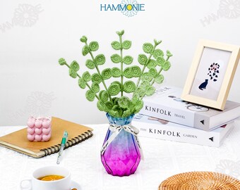 May include: A green crocheted plant in a pink and blue ombre glass vase. The vase is sitting on a white table with a book, a cup of coffee, and a notebook.