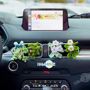 May include: Four crocheted flower air freshener car vent clips in a car. The flowers are in shades of green, white, and purple. The flowers are arranged in a row on the dashboard of a car. The text "HAMMONIE" is visible in the image.
