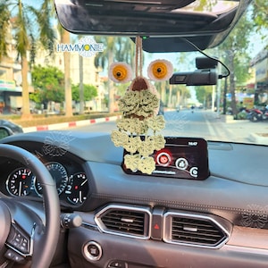 May include: A crocheted owl car charm with green and brown accents hanging from the rearview mirror of a car. The owl has large yellow eyes and a pink beak. The car's dashboard is visible in the background.