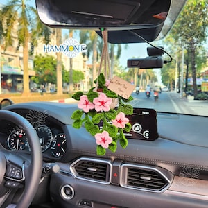 May include: A crocheted flower arrangement hanging from the rearview mirror of a car. The flowers are pink and white, and the leaves are green. A small wooden sign hangs from the arrangement with the text "I love you Mom & Dad".
