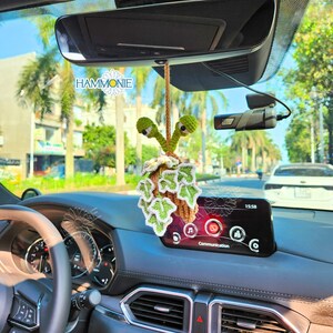 May include: A green and brown crocheted snail hanging from the rearview mirror of a car. The snail has large, googly eyes and is decorated with white flowers. The car's dashboard is visible in the background.