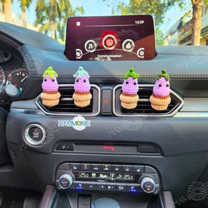May include: Four purple and green crocheted aubergine car air fresheners hanging from the air vents of a black car dashboard. The car has a digital display that reads "Communication" and "16:39".