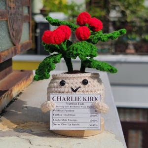 May include: A crocheted plant in a beige pot with a smiling face, holding a sign that reads "CHARLIE KIRK." The plant has green leaves and red berry-like accents. The sign lists "Nutrition Facts" and political attributes.