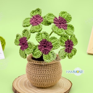 May include: A crocheted four-leaf clover plant in a brown pot. The plant is made of green and purple yarn and is sitting on a wooden base.