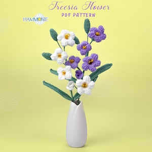 May include: A bouquet of crocheted freesia flowers in white and purple, arranged in a white vase on a yellow background. The flowers are arranged in a cluster with the stems extending down into the vase. The text "Freesia Flower PDF Pattern" is at the top of the image.