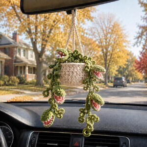 May include: A crocheted hanging plant ornament featuring a cream-colored pot and trailing green vines with multiple open-mouthed, toothy plant heads. The interior of each plant head is pink, and the entire piece is suspended by a braided cord.