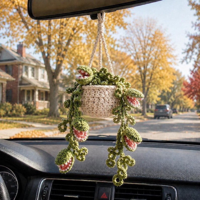 May include: A crocheted hanging plant ornament featuring a cream-colored pot and trailing green vines with multiple open-mouthed, toothy plant heads. The interior of each plant head is pink, and the entire piece is suspended by a braided cord.