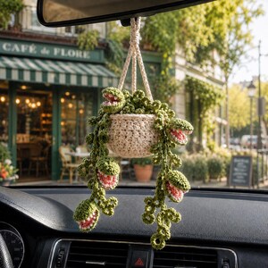 May include: A crocheted hanging plant ornament featuring a cream-colored pot and trailing green vines with multiple open-mouthed, pink-centered carnivorous plant heads. The decorative item is suspended by a braided cord, designed for car interior display.