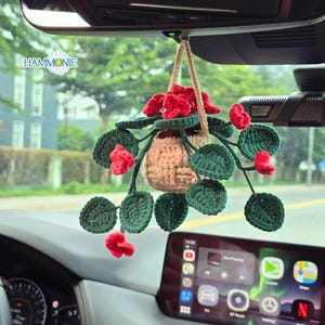 May include: A crocheted car charm depicting a hanging plant with red flowers and green leaves. The plant is in a beige pot, suspended by a beige string. The charm is hanging from a car's rearview mirror.