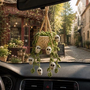 May include: A crocheted hanging plant ornament featuring a cream-colored pot and trailing green vines. Interspersed among the green leaves are several small, white, mummy-like figures with dark eyes, suspended by a braided cream-colored cord.