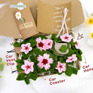 May include: A set of crocheted flower car accessories, including a hanging flower pot, a car vent clip, a car coaster, and a car hanging decoration. The flowers are pink and white with green leaves.