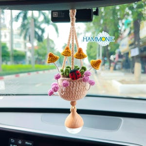 May include: A crocheted hanging planter with a pink basket and yellow mushroom accents. The planter is hanging from a rearview mirror in a car. The text "HAMMONIE" is visible in the background.