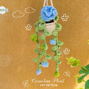 Op de afbeelding: Een gehaakte hangplant met blauwe bloemen en groene bladeren. De plant zit in een beige pot en hangt aan een touwtje. De achtergrond is een bruin, getextureerd oppervlak. De tekst "Carnation Plant PDF PATTERN" staat onderaan de afbeelding.
