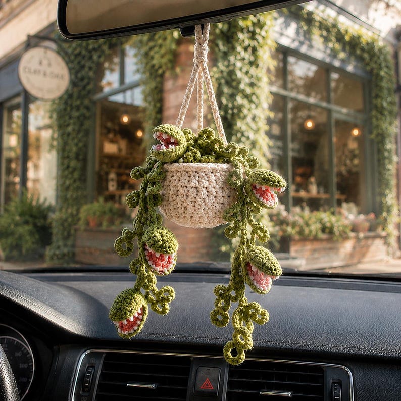 May include: A crocheted hanging plant ornament featuring a cream-colored pot and trailing green vines with multiple open-mouthed, toothy plant heads. The interior of each plant head is pink, and the entire piece is suspended by a braided cord.