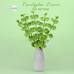 Puede incluir: Un arreglo de hojas de eucalipto de ganchillo en un jarrón blanco sobre un fondo verde claro. Las hojas son de un verde vibrante y el jarrón es de un blanco suave. El texto dice "Eucalyptus Leaves PDF Pattern".