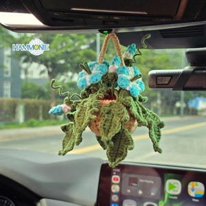 May include: A crocheted hanging plant decoration featuring blue and white flowers and green leaves. The plant is suspended from a car's rearview mirror. The word "HAMMONIE" is visible in the image.