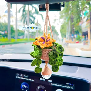 May include: A crocheted flower pot hanging from a rearview mirror in a car. The pot is filled with green crocheted leaves and a yellow and purple crocheted flower. The pot is hanging from a string with a crystal at the bottom.