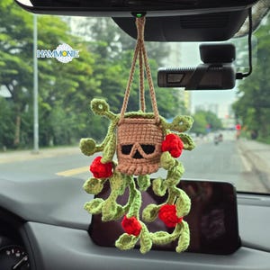 May include: Crocheted car charm featuring a skull design in a hanging basket. The charm is tan with a black skull, green vines, and red flowers. The charm is hanging from the rearview mirror.