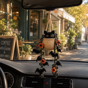 May include: A crocheted car accessory featuring a black cat with yellow eyes sitting in a beige pot. The cat is surrounded by hanging green vines decorated with small orange flowers and black bat shapes, suspended by a beige cord from a rearview mirror.