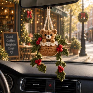 May include: A crocheted car hanging ornament featuring a brown bear peeking out of a tan basket, accented with green holly leaves and red berries. The bear has black eyes and a stitched nose. It is suspended by a cream-colored braided cord.