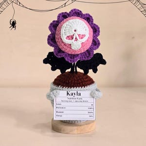 Peut inclure: Une sculpture de fleur au crochet avec un centre de cr&acirc;ne, des p&eacute;tales violets et des accents de chauve-souris noirs. La sculpture a une base marron et un pot bleu clair. Une pancarte indique "Kayla" et "Nutrition Facts".