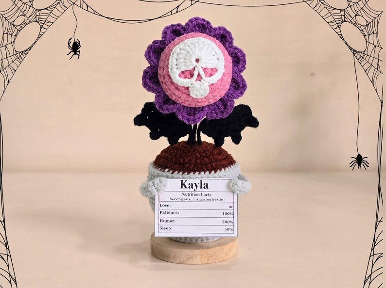 Peut inclure: Une sculpture de fleur au crochet avec un centre de cr&acirc;ne, des p&eacute;tales violets et des accents de chauve-souris noirs. La sculpture a une base marron et un pot bleu clair. Une pancarte indique "Kayla" et "Nutrition Facts".