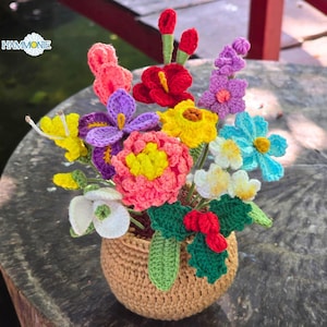 May include: A handcrafted crochet flower arrangement in a small, woven basket. The bouquet features a variety of colorful flowers, including red, pink, yellow, purple, and blue blooms. The arrangement is set on a rustic wooden surface. The word "HAMMONIE" is in the top left corner.