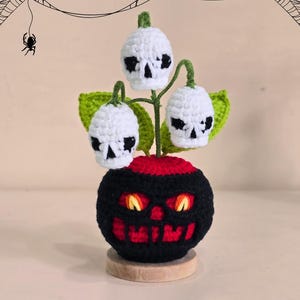 Peut inclure: D&eacute;coration d'Halloween en crochet repr&eacute;sentant une base noire et rouge en forme de citrouille avec des yeux orange brillants et une bouche souriante. Trois fleurs blanches en forme de cr&acirc;ne avec des feuilles vertes sortent du dessus.